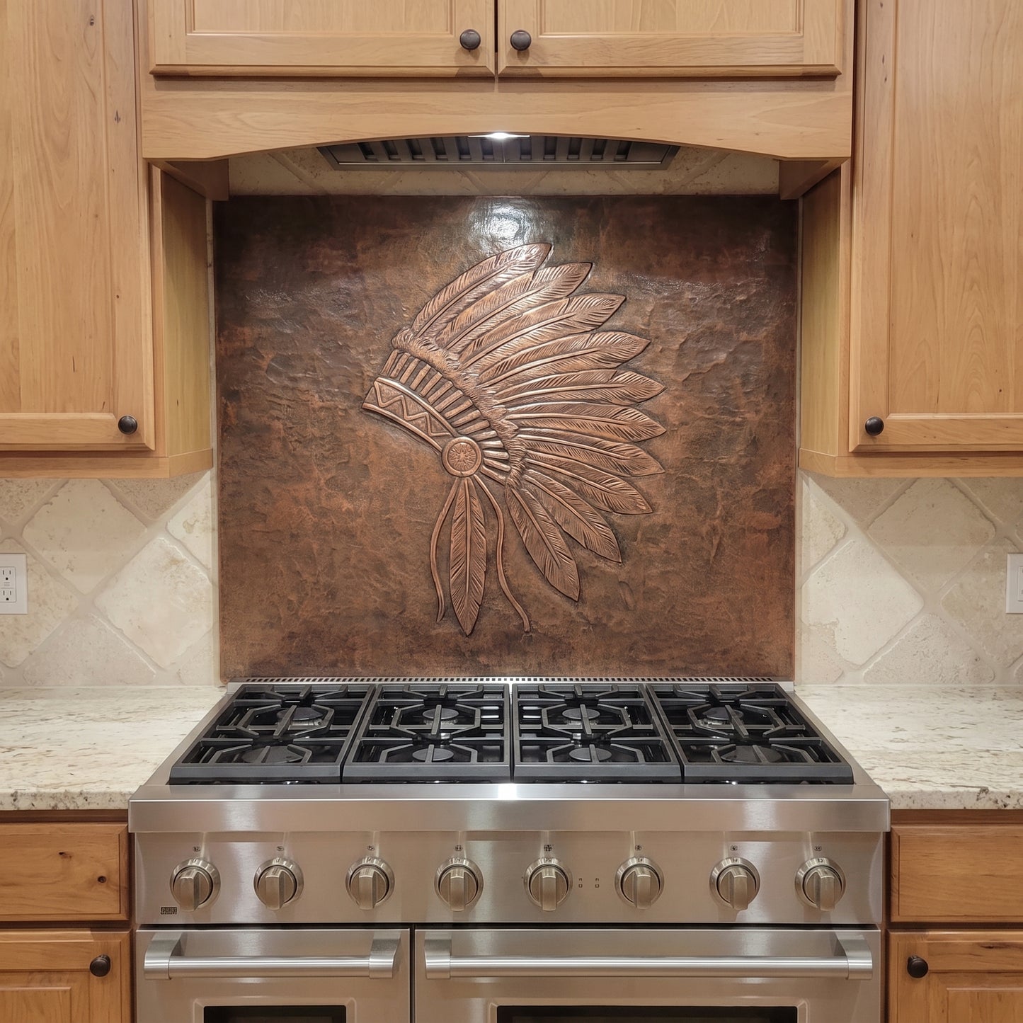 Handcrafted feather headdress copper backsplash installed behind a stainless steel range, featuring a large raised-relief feather design on a hammered copper background in a warm wood kitchen.