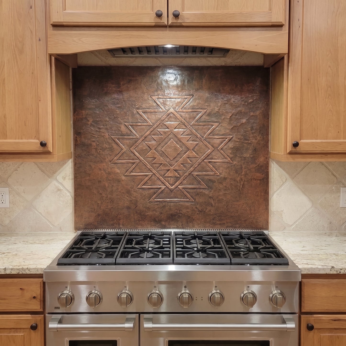 Handcrafted copper stove backsplash installed behind a stainless steel range, featuring a large Southwestern geometric diamond pattern in raised relief centered on a richly hand-hammered copper background, with natural wood cabinets and light stone countertops surrounding the panel.