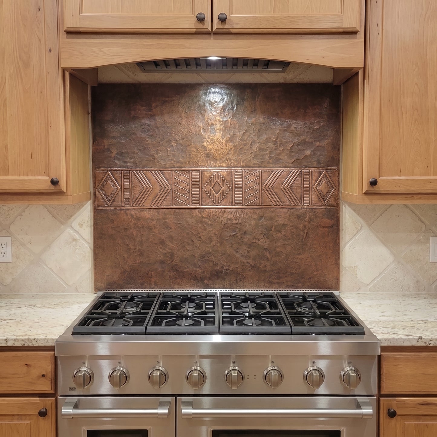 Handcrafted Southwestern copper stove backsplash installed behind a stainless steel range, featuring a wide raised geometric band with diamonds, chevrons, stepped lines, and repeating linear patterns across the center of a hand-hammered brown copper panel. The custom copper kitchen backsplash is shown between natural wood cabinets with light stone countertops and surrounding tile.
