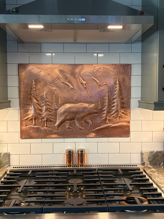 A rectangular copper relief wall art piece depicting a bear, trees, and mountains, mounted on a white subway tile backsplash above a gas range.