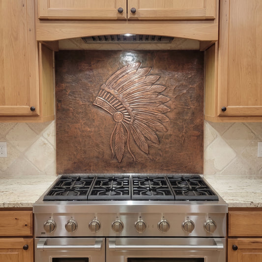 Handcrafted feather headdress copper backsplash installed behind a stainless steel range, featuring a large raised-relief feather design on a hammered copper background in a warm wood kitchen.