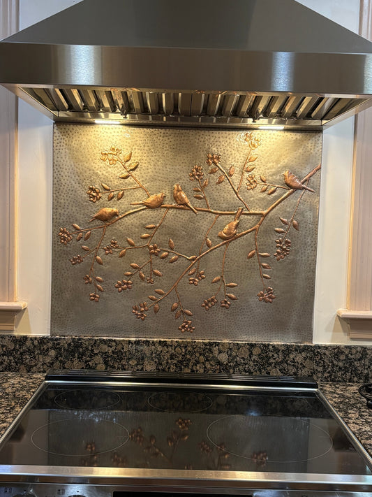 A decorative hammered metal backsplash panel with an embossed branch and bird design, installed behind a stainless steel range hood and above a black glass cooktop in a kitchen.