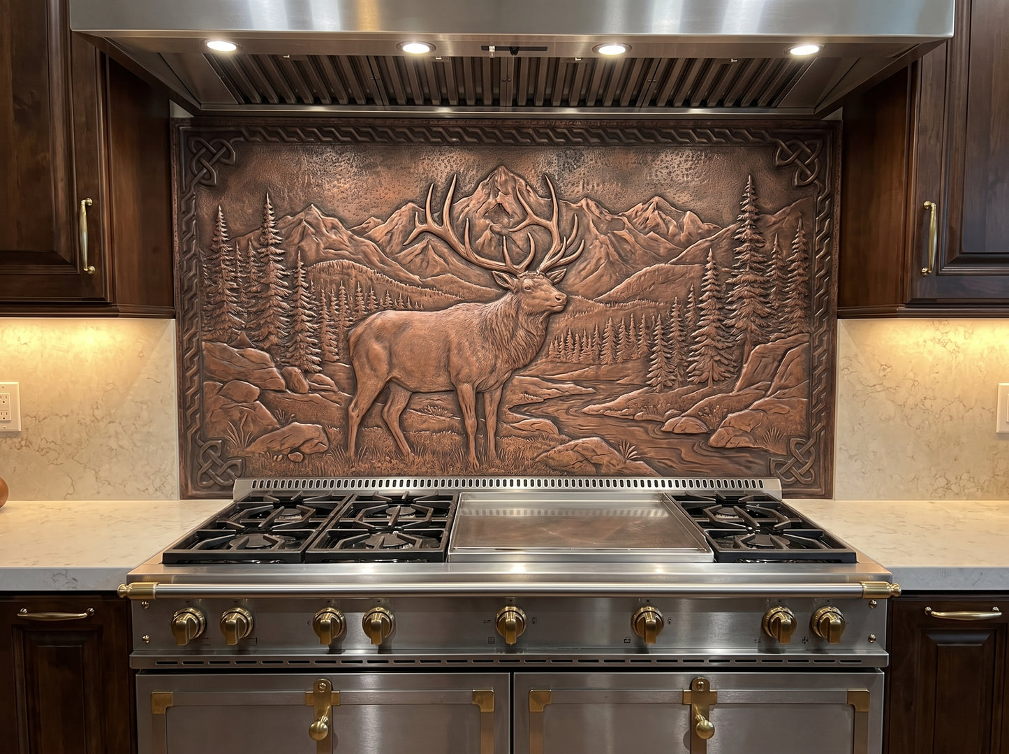 Rocky Mountain Elk copper kitchen backsplash installed behind a professional stainless steel range, with a large bull elk, detailed antlers, pine forest, mountain landscape, rocky terrain, and flowing stream handcrafted in raised metal relief.