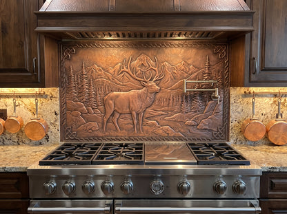 Handcrafted Rocky Mountain Elk copper stove backsplash installed behind a large range with a wall-mounted pot filler, featuring a dimensional bull elk against mountain peaks, evergreen trees, rocks, and a winding woodland stream.
