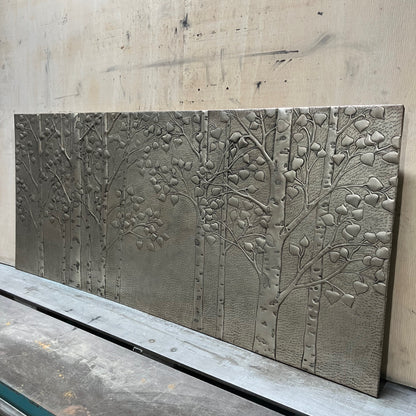 A rectangular metal wall art panel featuring embossed tree branches and leaves, displayed on a workbench against a light wood background.