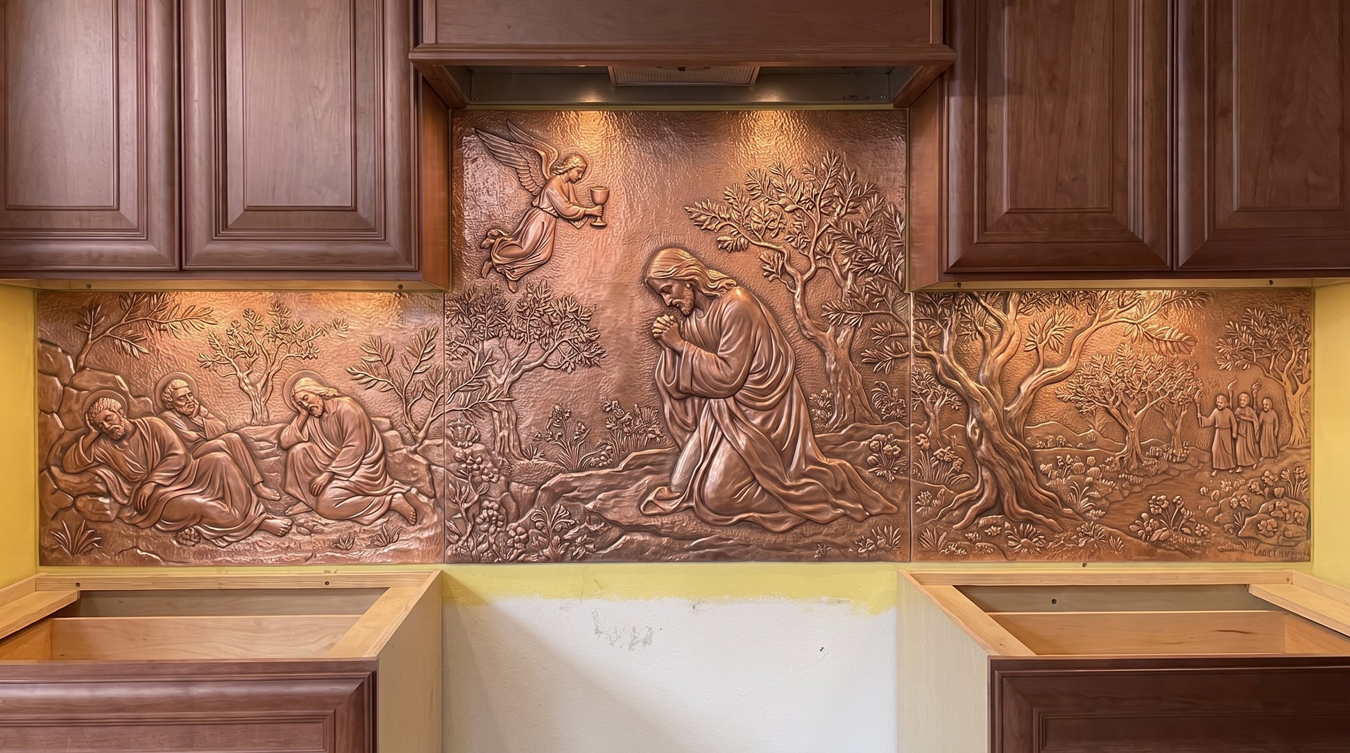 Agony in the Garden copper kitchen backsplash depicting Jesus praying in the Garden of Gethsemane, with disciples, approaching figures, olive trees, garden vegetation, and landscape details telling the biblical Gethsemane story in raised metal relief across the range wall.