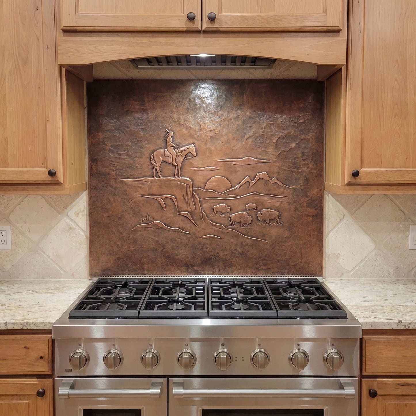 Handcrafted copper stove backsplash installed behind a stainless steel range, featuring a Native American rider on horseback overlooking a small herd of buffalo in raised relief, with distant mountains, a setting sun, rocky Western terrain, and a richly hand-hammered copper background.