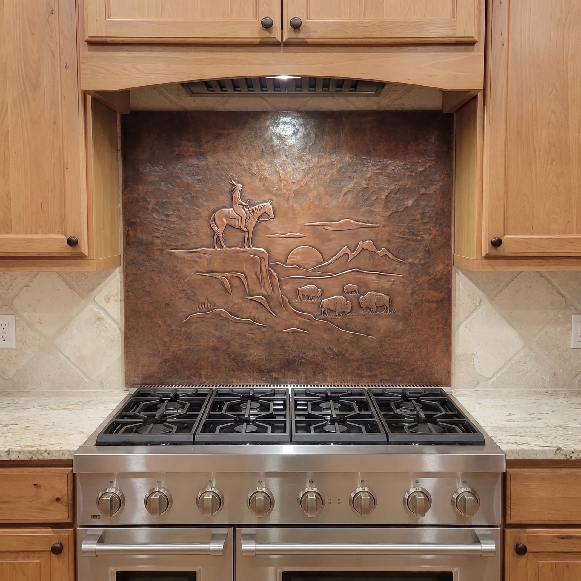 Handcrafted copper stove backsplash installed behind a stainless steel range, featuring a Native American rider on horseback overlooking a small herd of buffalo in raised relief, with distant mountains, a setting sun, rocky Western terrain, and a richly hand-hammered copper background.