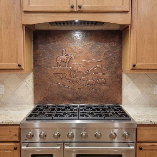 Handcrafted copper stove backsplash installed behind a stainless steel range, featuring a Native American rider on horseback overlooking a small herd of buffalo in raised relief, with distant mountains, a setting sun, rocky Western terrain, and a richly hand-hammered copper background.