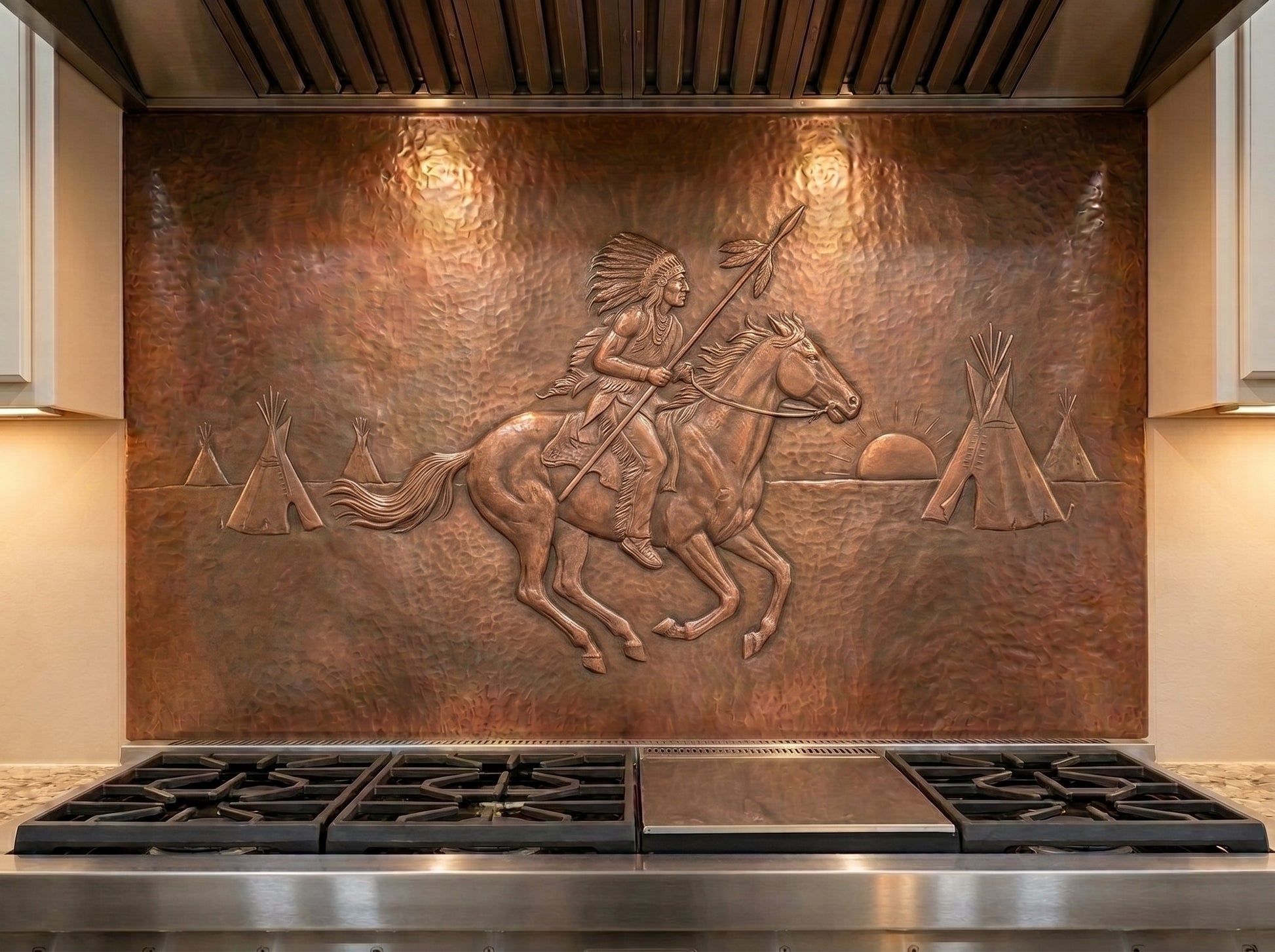 Handcrafted copper stove backsplash installed behind a professional range, featuring a Native American rider on horseback holding a spear in raised relief, with a feather headdress, tipis, setting sun, and hand-hammered copper background in a Western kitchen.