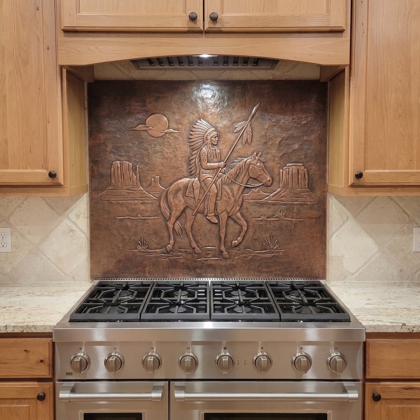 Handcrafted copper stove backsplash installed behind a stainless steel range, featuring a Native American-inspired horseback warrior holding a feathered spear in raised relief, with desert mesas, a setting sun, and sparse Southwestern landscape details formed against a hand-hammered copper background.