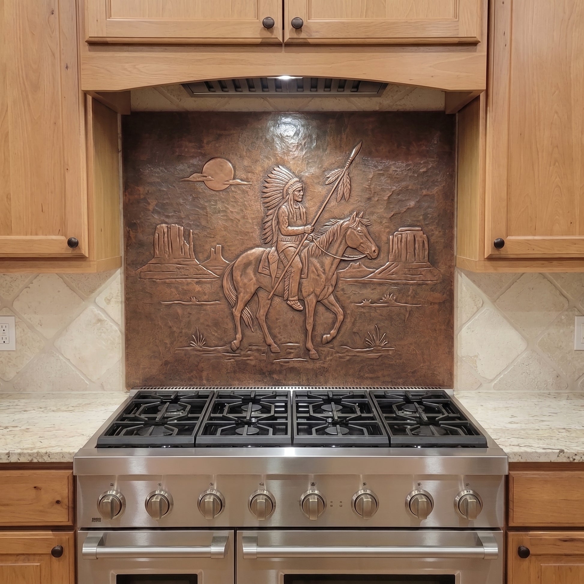 Handcrafted copper stove backsplash installed behind a stainless steel range, featuring a Native American-inspired horseback warrior holding a feathered spear in raised relief, with desert mesas, a setting sun, and sparse Southwestern landscape details formed against a hand-hammered copper background.