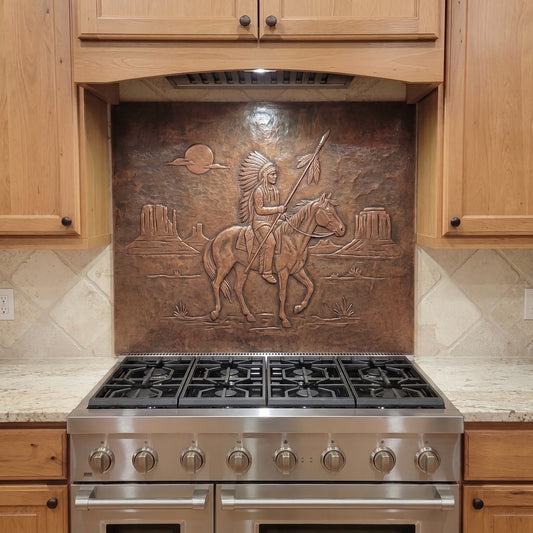 Handcrafted copper stove backsplash installed behind a stainless steel range, featuring a Native American-inspired horseback warrior holding a feathered spear in raised relief, with desert mesas, a setting sun, and sparse Southwestern landscape details formed against a hand-hammered copper background.