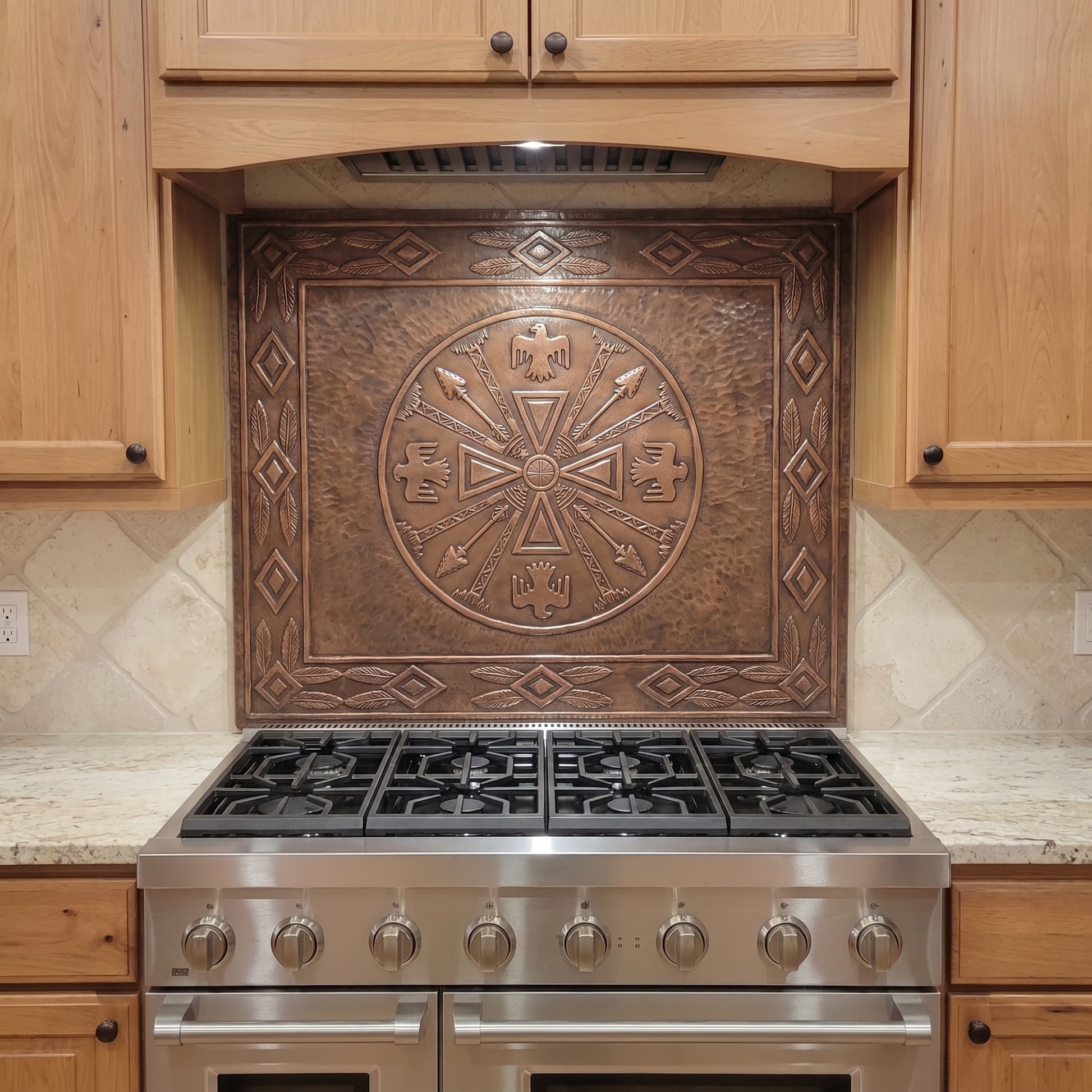 Handcrafted copper stove backsplash installed behind a stainless steel range, featuring a large Native American-inspired medicine wheel in raised relief with eagles, crossed arrows, feathers, geometric symbols, and a detailed decorative border on a hand-hammered copper background.