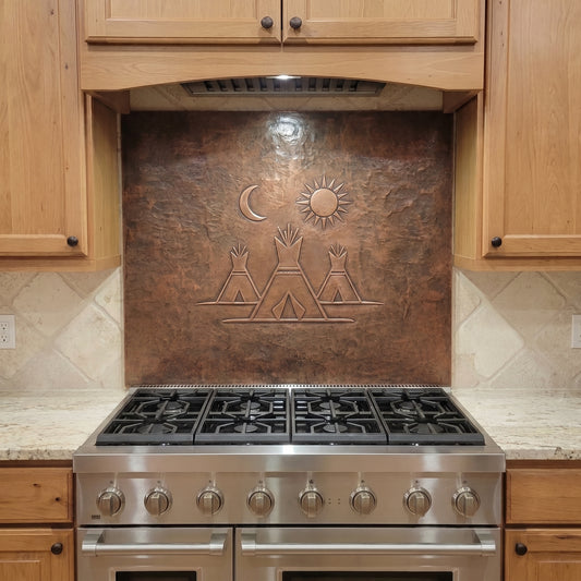 Handcrafted copper stove backsplash installed behind a stainless steel range, featuring three Native American-inspired teepees beneath a raised sun and crescent moon. The simple Southwestern design is formed in raised relief against a hand-hammered brown copper background and shown between natural wood kitchen cabinets.