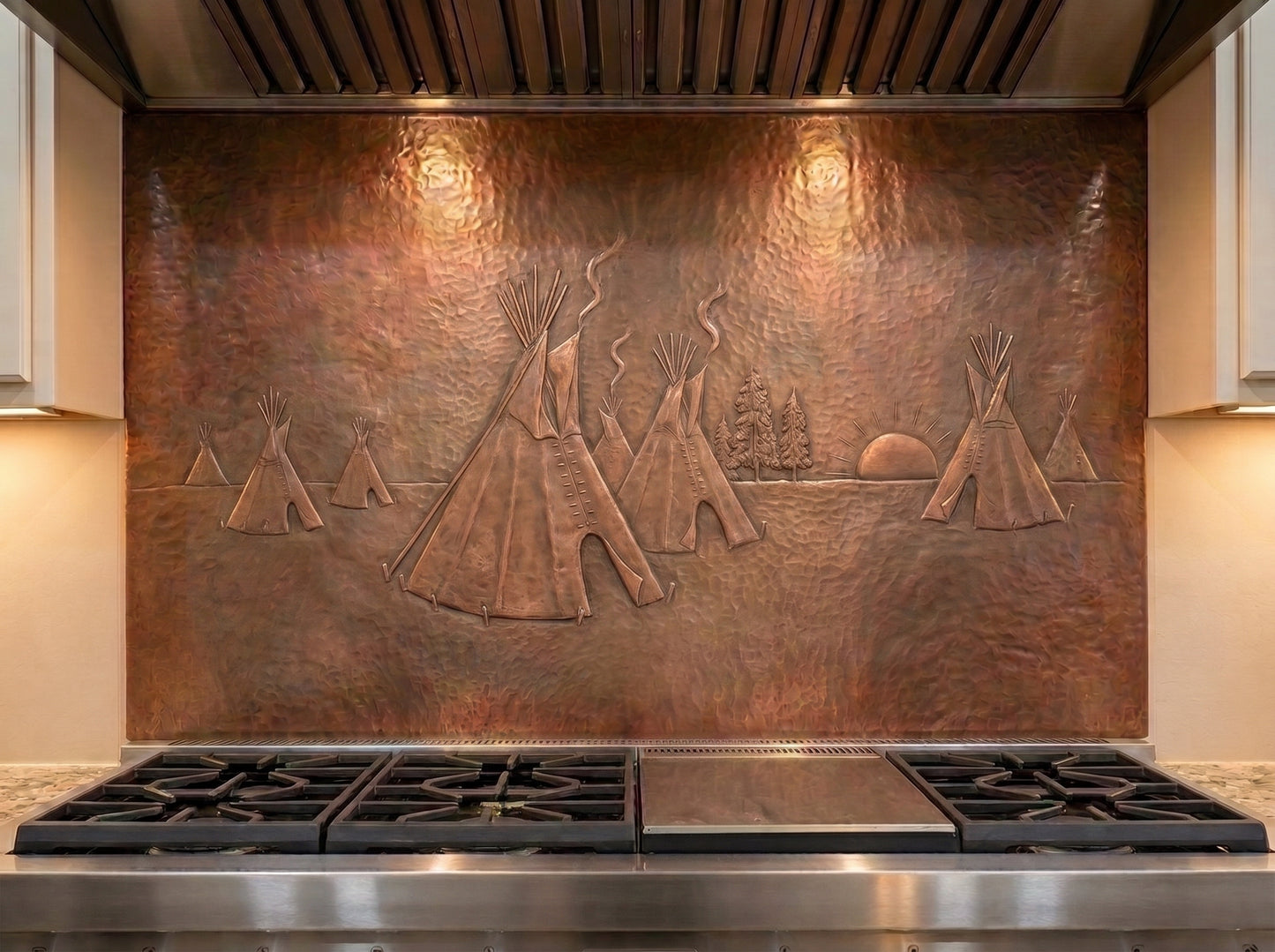 Handcrafted copper stove backsplash installed behind a professional gas range, featuring a Native American tipi village in raised relief with several tipis, smoke rising from the camp, pine trees, and a setting sun across a richly hand-hammered copper background.