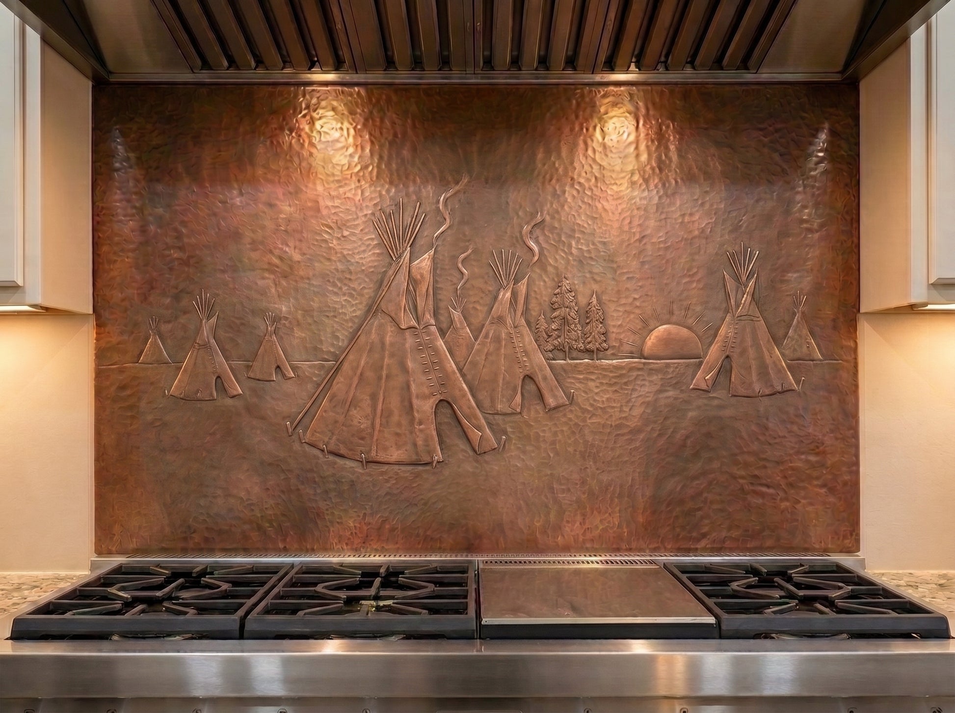 Handcrafted copper stove backsplash installed behind a professional gas range, featuring a Native American tipi village in raised relief with several tipis, smoke rising from the camp, pine trees, and a setting sun across a richly hand-hammered copper background.