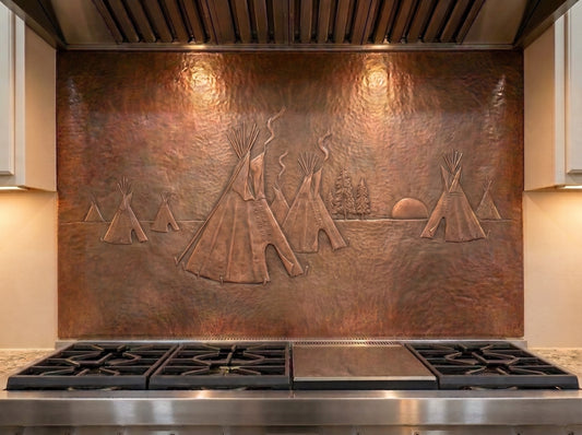Handcrafted copper stove backsplash installed behind a professional gas range, featuring a Native American tipi village in raised relief with several tipis, smoke rising from the camp, pine trees, and a setting sun across a richly hand-hammered copper background.