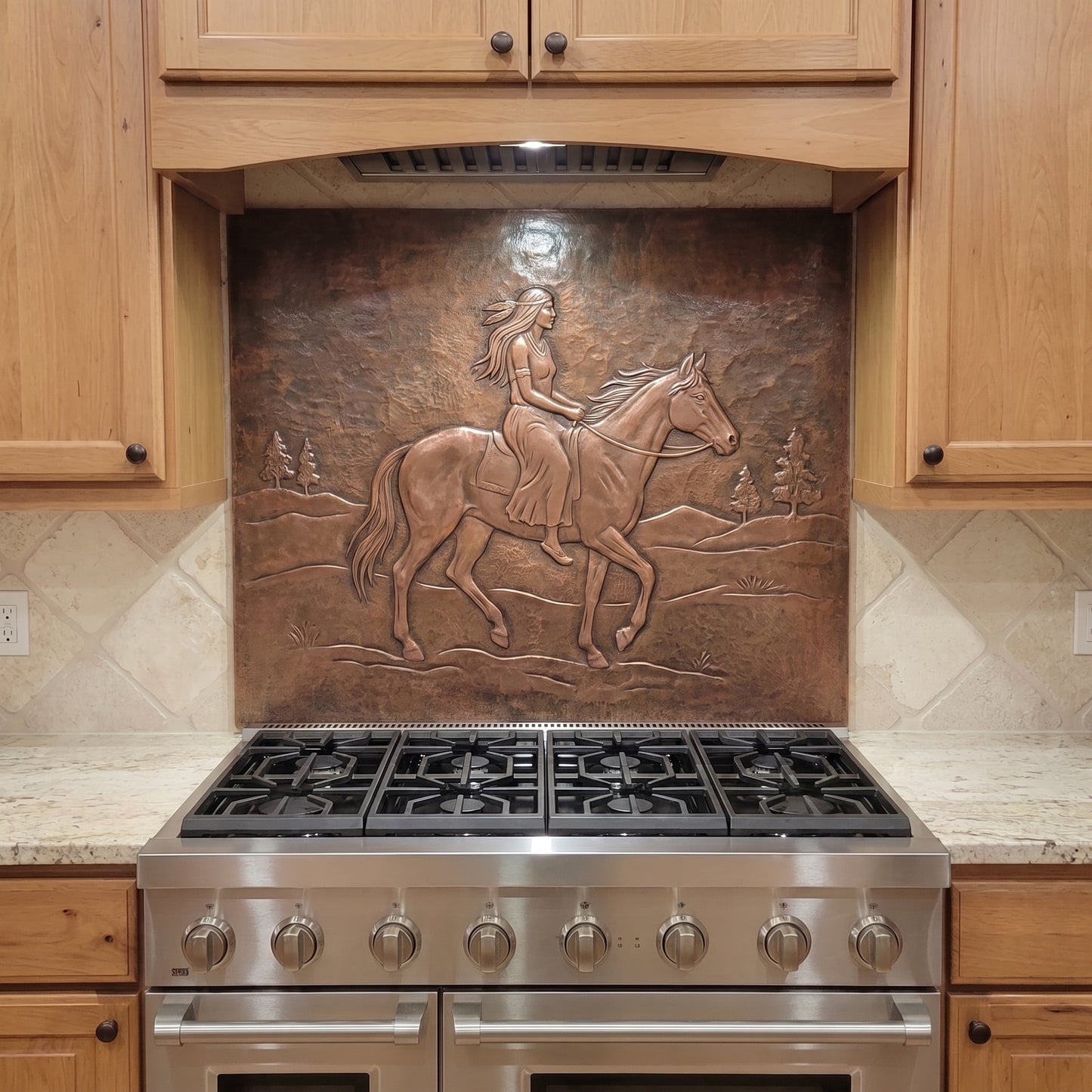 Handcrafted copper stove backsplash installed behind a stainless steel range, featuring a Native woman riding a horse in raised relief across an open Western landscape with distant hills and pine trees, surrounded by natural wood kitchen cabinets and light stone countertops.