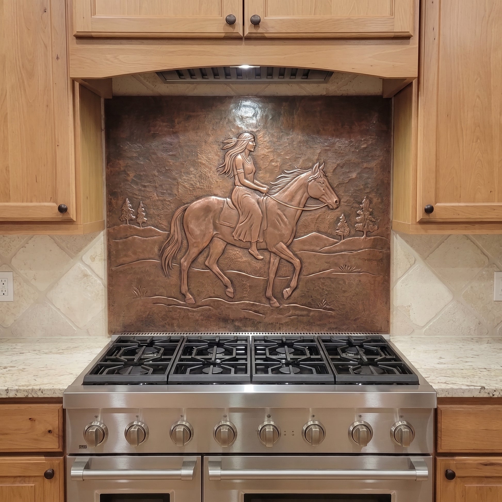 Handcrafted copper stove backsplash installed behind a stainless steel range, featuring a Native woman riding a horse in raised relief across an open Western landscape with distant hills and pine trees, surrounded by natural wood kitchen cabinets and light stone countertops.