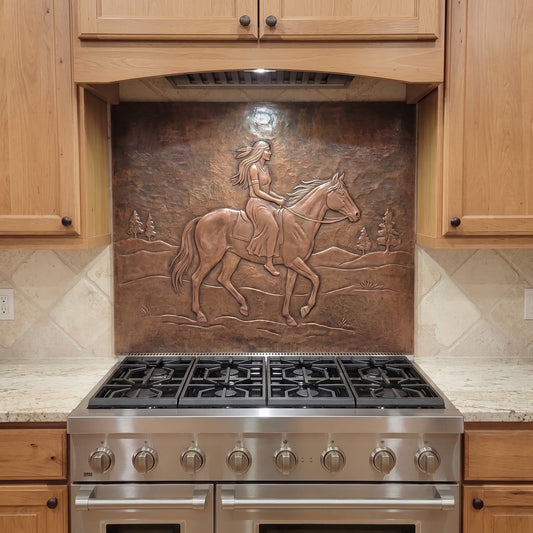 Handcrafted copper stove backsplash installed behind a stainless steel range, featuring a Native woman riding a horse in raised relief across an open Western landscape with distant hills and pine trees, surrounded by natural wood kitchen cabinets and light stone countertops.