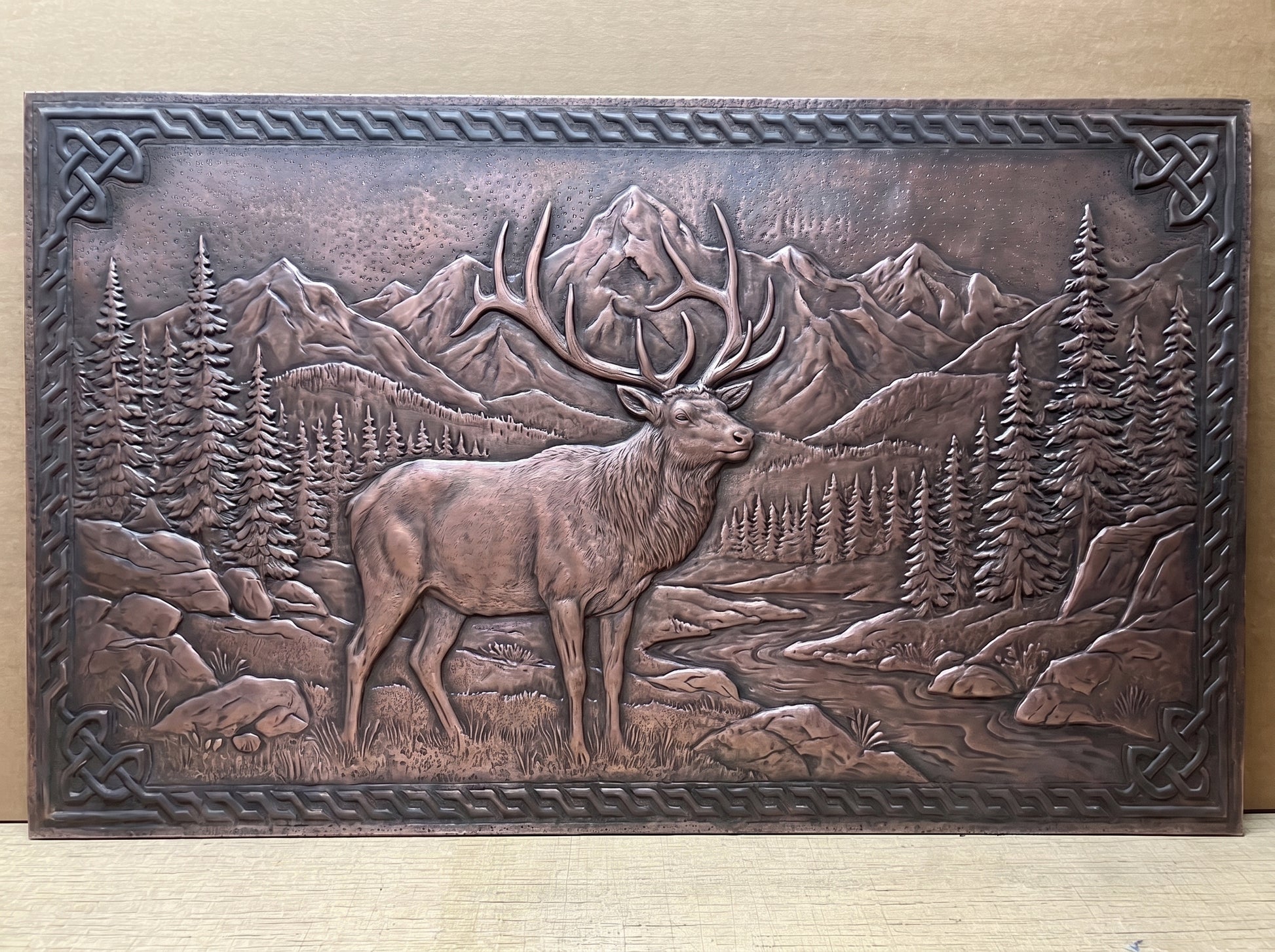 Rocky Mountain Elk handcrafted copper panel featuring a bull elk with large detailed antlers standing in a mountain landscape with pine forests, rocky terrain, distant peaks, and a winding stream, surrounded by a raised Celtic-inspired decorative border.