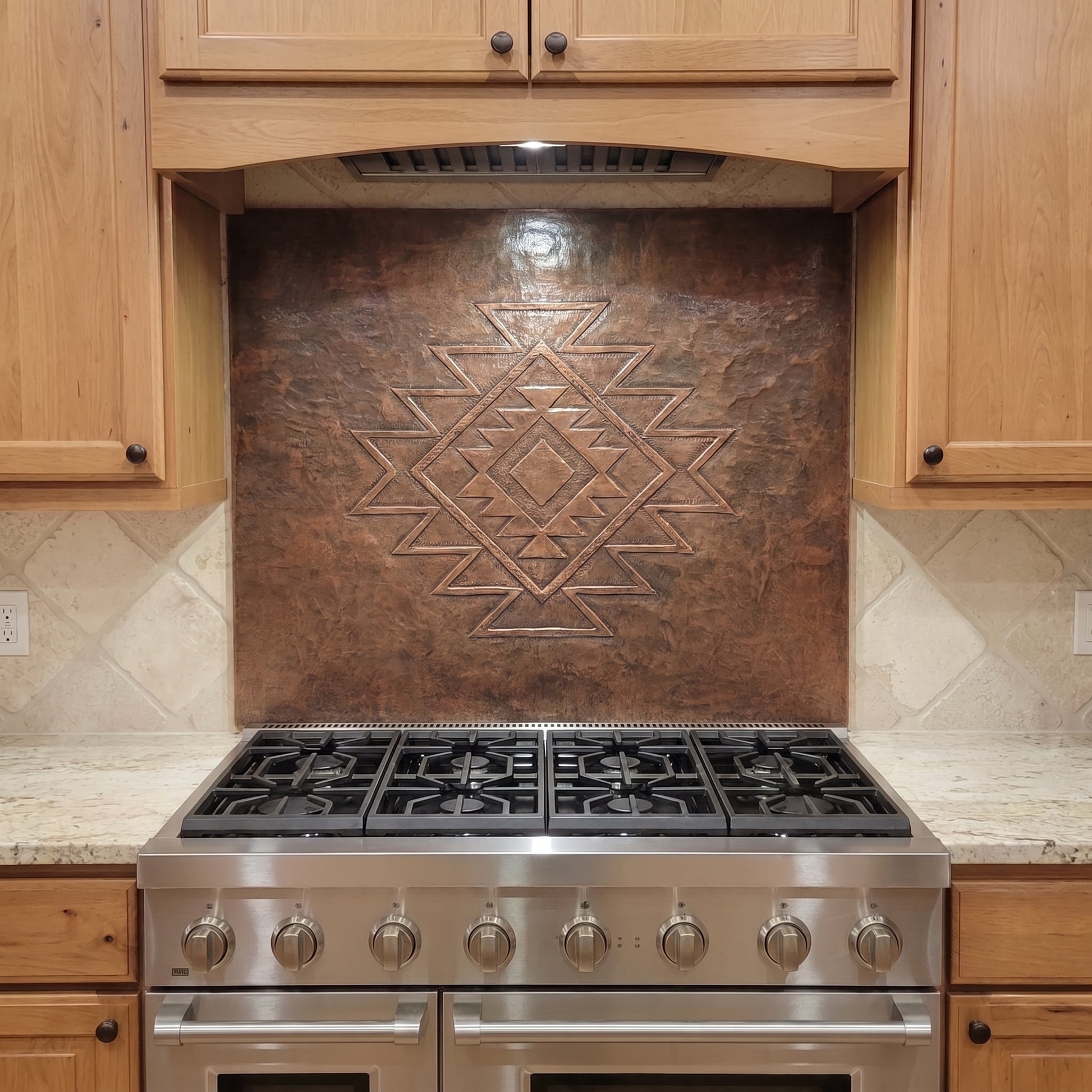 Handcrafted copper stove backsplash installed behind a stainless steel range, featuring a large Southwestern geometric diamond pattern in raised relief centered on a richly hand-hammered copper background, with natural wood cabinets and light stone countertops surrounding the panel.