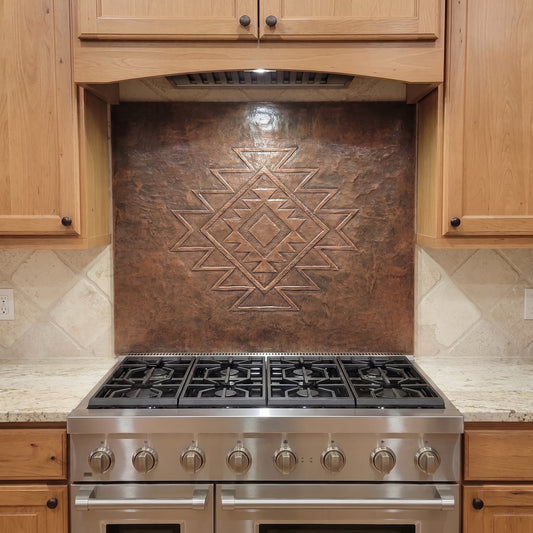 Handcrafted copper stove backsplash installed behind a stainless steel range, featuring a large Southwestern geometric diamond pattern in raised relief centered on a richly hand-hammered copper background, with natural wood cabinets and light stone countertops surrounding the panel.