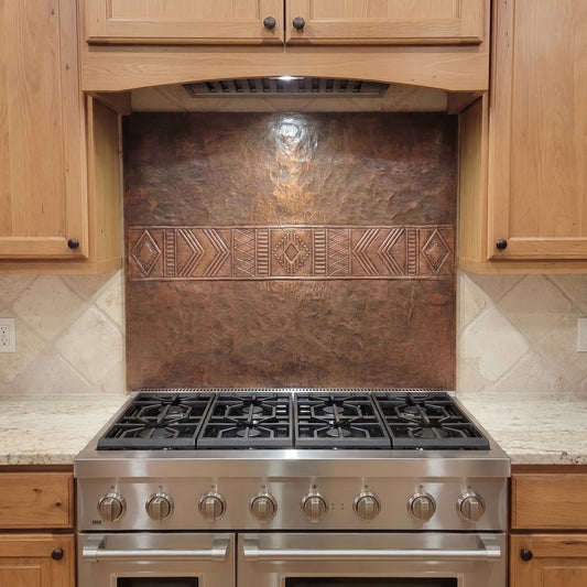 Handcrafted Southwestern copper stove backsplash installed behind a stainless steel range, featuring a wide raised geometric band with diamonds, chevrons, stepped lines, and repeating linear patterns across the center of a hand-hammered brown copper panel. The custom copper kitchen backsplash is shown between natural wood cabinets with light stone countertops and surrounding tile.