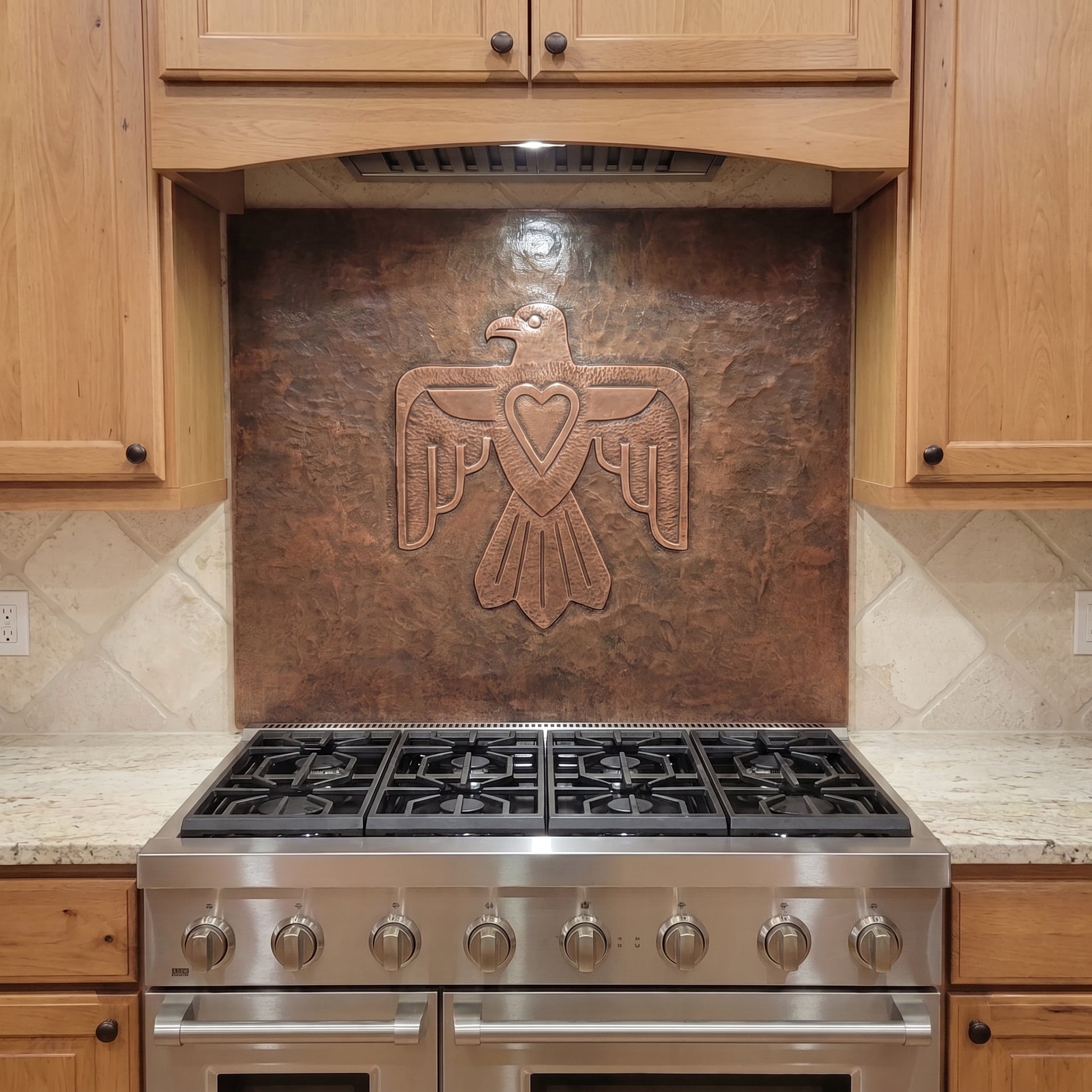 Handcrafted Thunderbird copper backsplash installed behind a stainless steel range, featuring a large raised Thunderbird with outstretched wings and a heart at its center on a hand-hammered metal background, surrounded by warm natural wood kitchen cabinets.