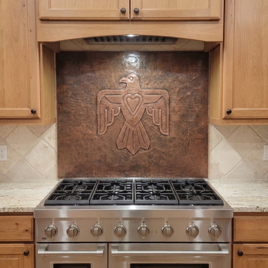 Handcrafted Thunderbird copper backsplash installed behind a stainless steel range, featuring a large raised Thunderbird with outstretched wings and a heart at its center on a hand-hammered metal background, surrounded by warm natural wood kitchen cabinets.