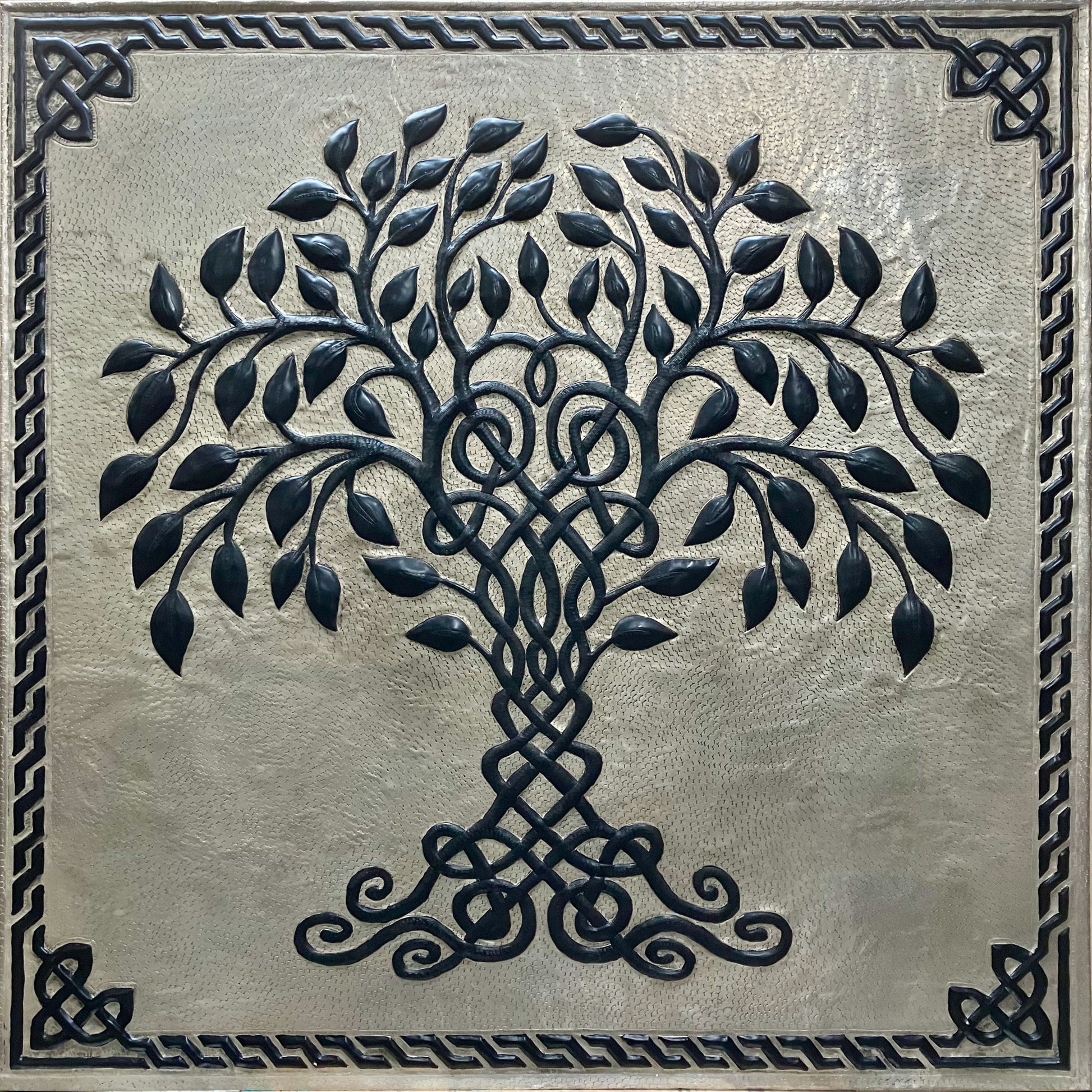 A square decorative wall plaque featuring a raised black tree of life with intricate Celtic knotwork in the trunk and branches, set against a textured metallic silver background with a matching Celtic border.