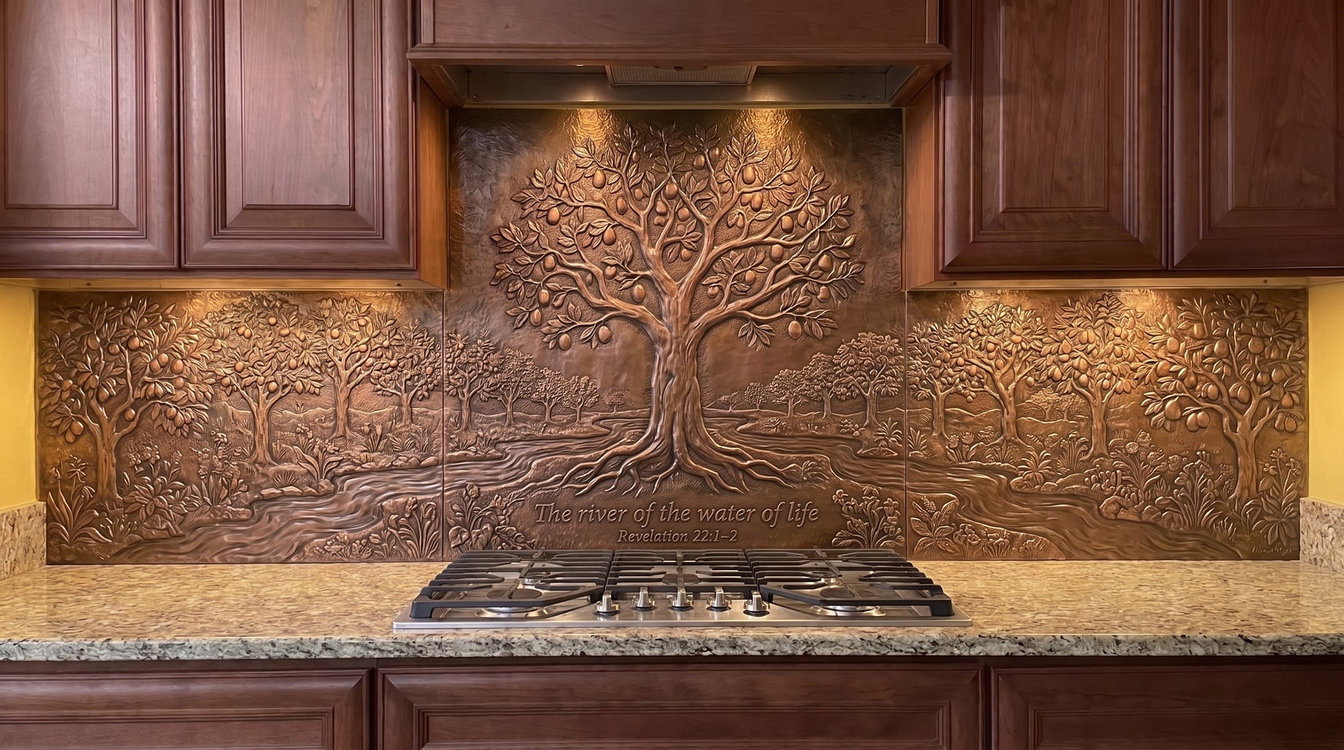 Handcrafted Tree of Life and Living Waters copper kitchen backsplash inspired by Revelation 22:1–2, featuring a large Tree of Life beside flowing water with abundant trees and landscape details extending across the backsplash behind the range and under the kitchen cabinets.