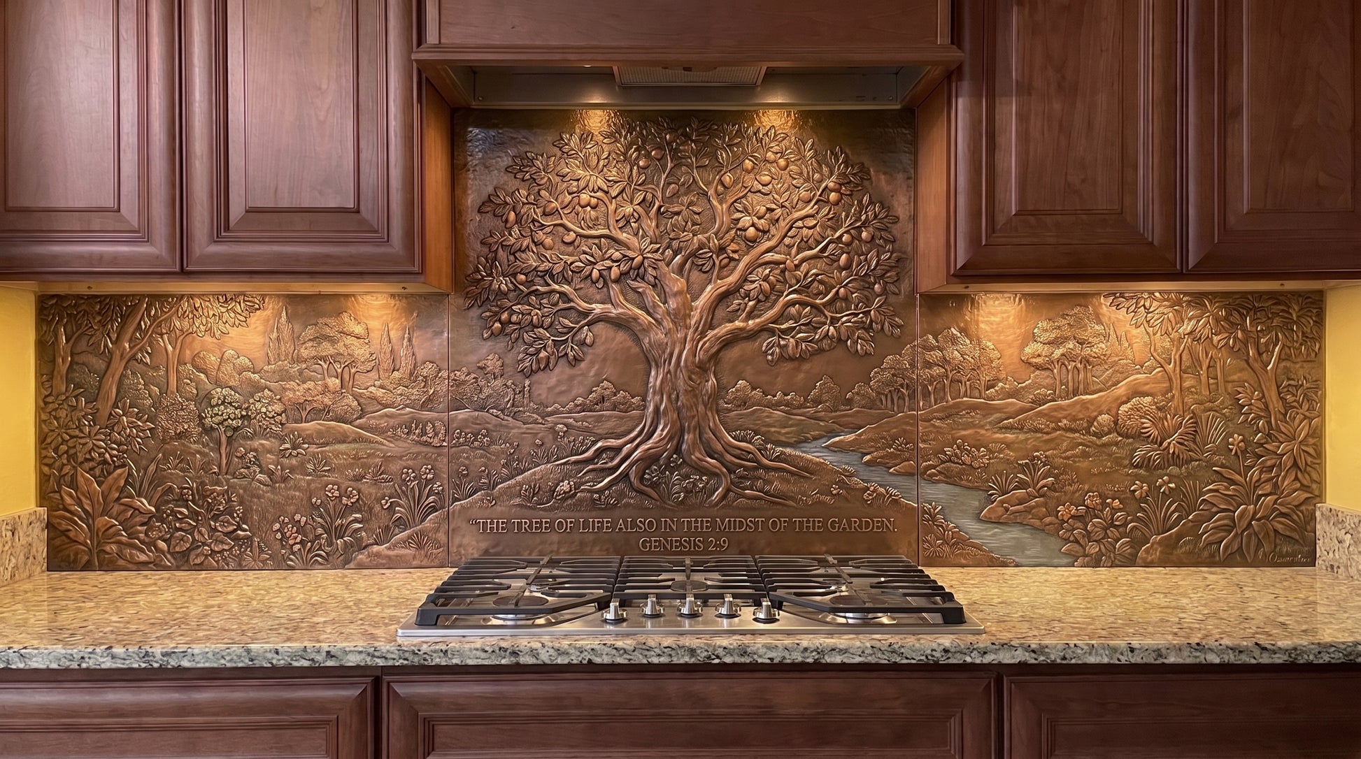 Handcrafted Tree of Life in Eden copper kitchen backsplash inspired by Genesis 2:9, featuring a large Tree of Life in a lush Garden of Eden landscape with dense vegetation, smaller trees, rolling ground, and a stream extending across the copper backsplash behind the range and under the cabinets.
