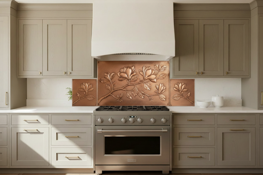 Three copper panels spanning full kitchen wall behind 36-inch range — graceful magnolia branch with large open blooms, closed buds and oval leaves in repoussé relief across all three panels, warm natural copper tone matching magnolia reference, fine stipple background, warm greige shaker cabinets with brushed gold hardware, white quartz countertop, transitional kitchen